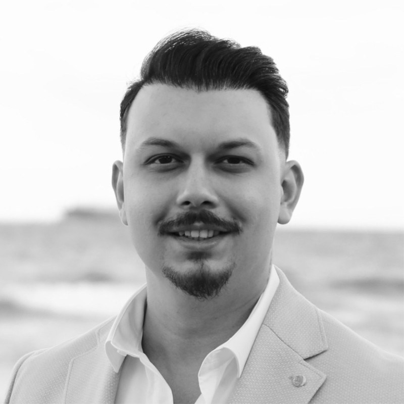 Portrait of Berk Atayman in a light blazer and white shirt, smiling, with the sea behind him